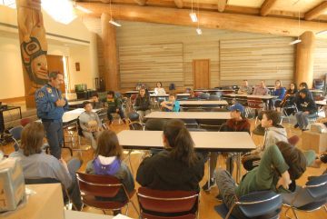 Youth are sitting in chairs in a circle in a wooden hall with. Astronaut John Herrington is standing at the edge of the circle wearing a blue NASA bomber jacket.